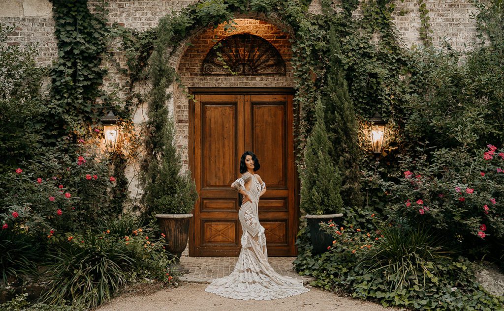 Bride in a boho lace gown standing in front of a wooden door and vines at Hidden Waters Dallas Wedding Venue