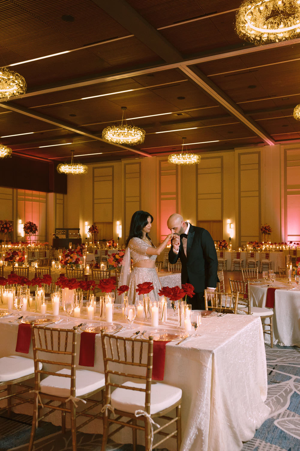 Indian b ride and groom in their wedding reception hall. Groom is kissing the bride's hand, while they are surrounded by romantic candles and red roses in their Texas wedding venue, the Omni PGA Dallas. Romantic, low-light, an image that looks quiet and full of adoration.