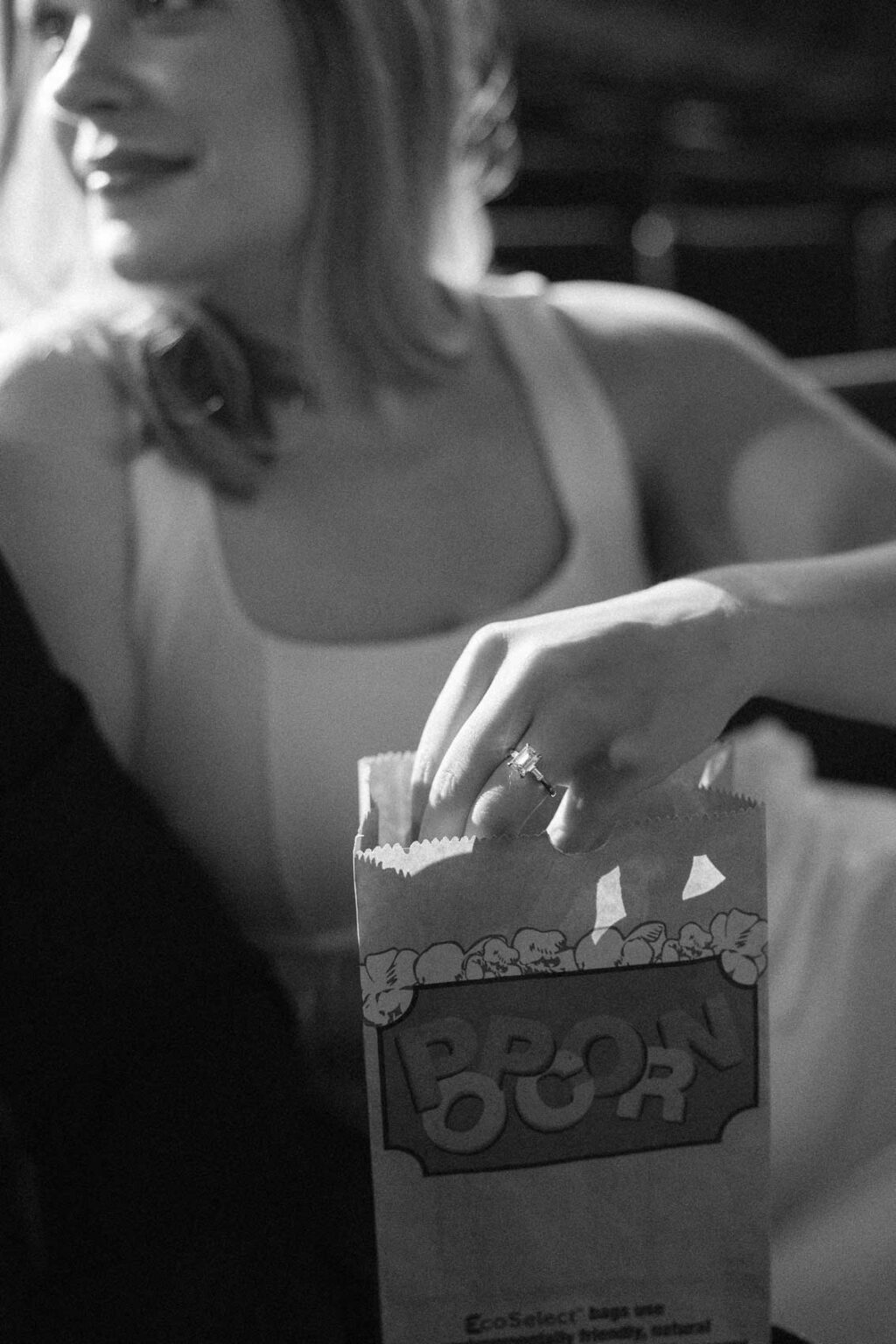 Black & white photo od Ginny and Deva with her hand inside a bag of popcorn, her engagement ring flashing in the light, SIFF Cinema Uptown Seattle, dramatic theater lighting, film-style engagement photography