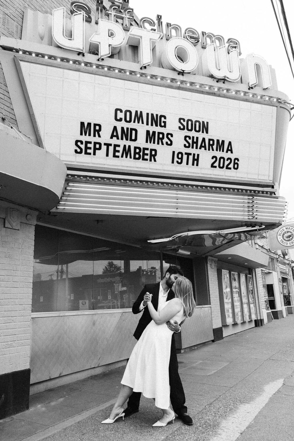 Black & white photo of a couple standing beneath a lit movie theater marquee sign with their wedding date, SIFF Cinema Uptown Seattle